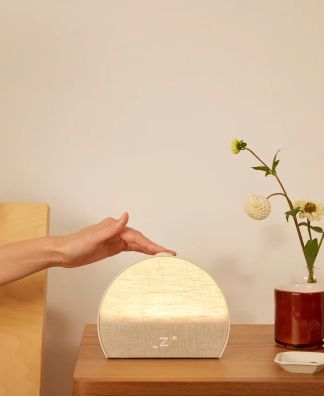 Hand tapping the top button on a Hatch Restore 3 sitting on a nightstand